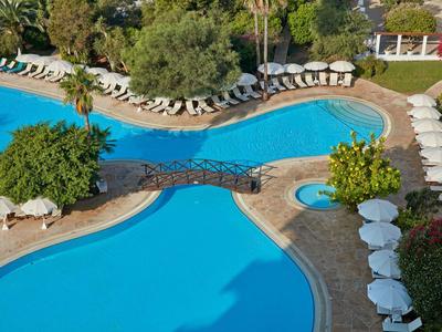 Großer, blauer Hotelpool mit Steg, umgeben von weißen Sonnenschirmen und grüner Vegetation.