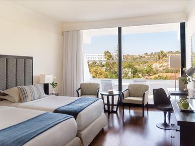 Modernes Hotelzimmer mit zwei Einzelbetten, Sitzbereich und Balkon mit Blick auf grüne Hügel.