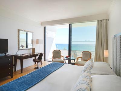 Helles Hotelzimmer mit Meerblick, Doppelbett, Schreibtisch, Stühlen und großem Fenster.