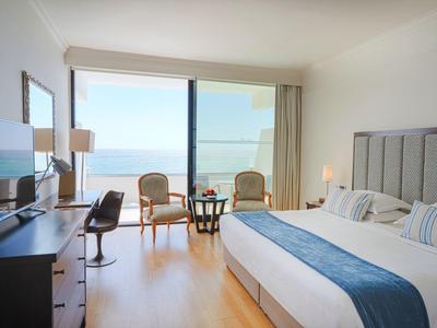 Hell eingerichtetes Hotelzimmer mit Meerblick, Doppelbett und Sitzbereich am Fenster.