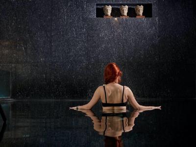 Frau mit roten Haaren entspannt im Innenpool unter sanftem Regenlicht in dunklem Raum.