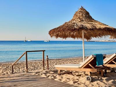 Sandstrand mit Strohschirm, Liegen und Blick auf ruhiges, blaues Meer bei klarem Himmel.
