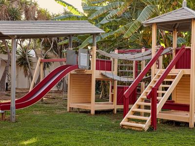 Spielplatz im Hotelgarten mit zwei Rutschen, Holzhäusern und grüner Wiese.