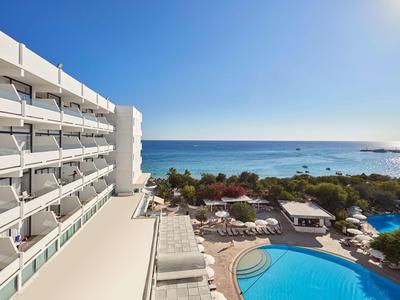 Hotel mit rundem Pool, Sonnenliegen und Blick auf das Meer bei klarem Himmel.
