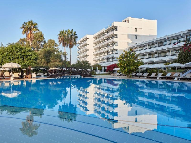 Großer Pool mit Sonnenliegen vor einem mehrstöckigen Hotelgebäude unter blauem Himmel.