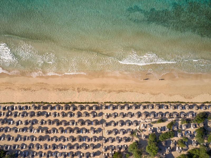 Luftaufnahme eines Strandes mit Reihen von Sonnenschirmen und klarem Wasser.