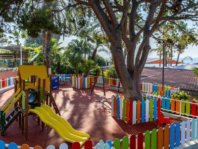Bunter Spielplatz mit Rutsche, Schaukeln und Bäumen, umgeben von einem farbigen Zaun.