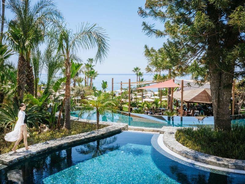 Venkovní hotelový bazén s palmami, slunečními plachtami a uvolněnou atmosférou.