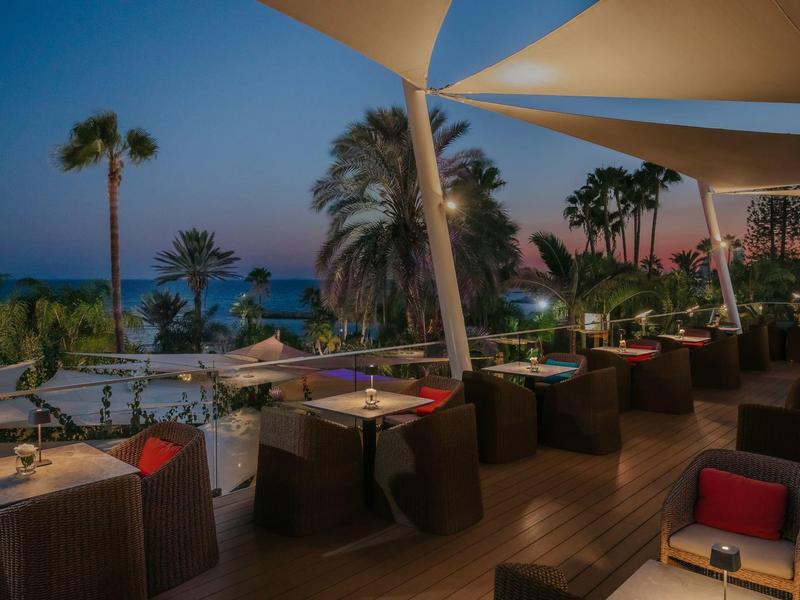 Večerní terasa s posezením a výhledem na palmy a moře při západu slunce.