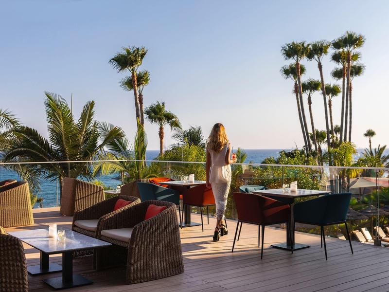 Žena v bílých šatech na terase s palmami a výhledem na oceán při západu slunce