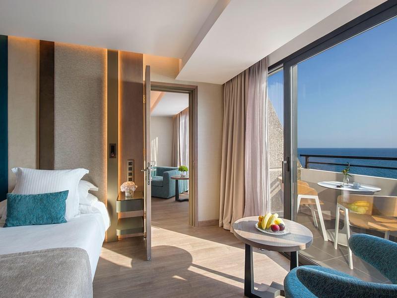 Moderní hotelový pokoj s postelí, posezením a balkonem s výhledem na moře za denního světla.