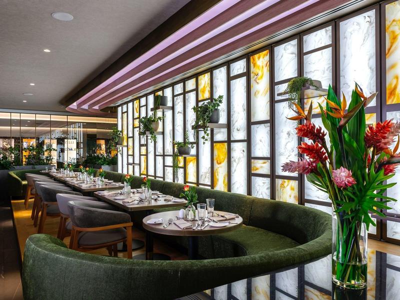 Moderní restaurace s dlouhými zelenými pohovkami, kulatými stoly a rostlinami u osvětlených oken.