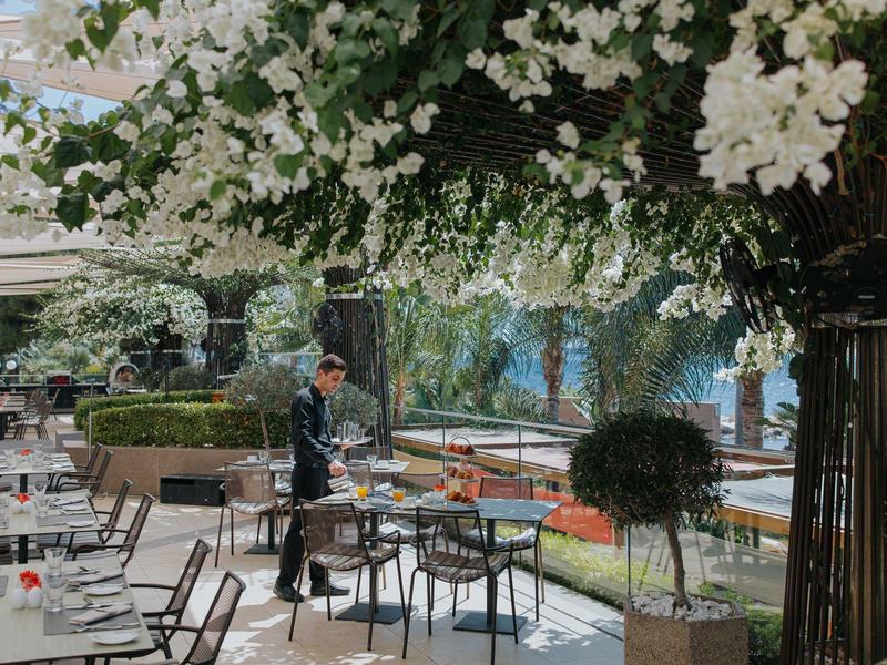 Venkovní terasa restaurace pokrytá květinami, se stoly, židlemi a prostřením za denního světla.