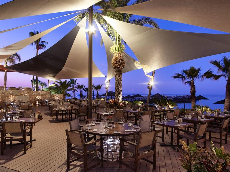 Otevřená restaurace na pláži během západu slunce se dřevěnými židlemi a ubrusy pod plachtovými stany.