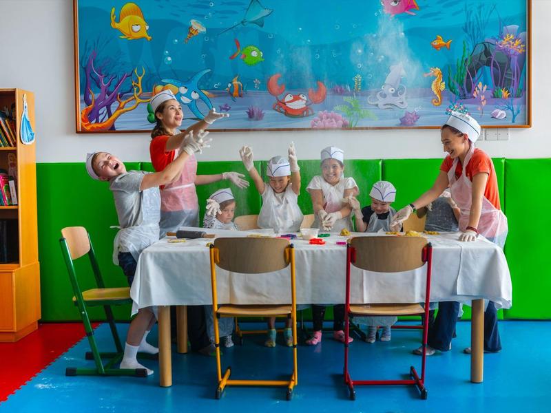 Děti v kuchařských čepicích a zástěrách pečou u prostřeného stolu v barevné místnosti.