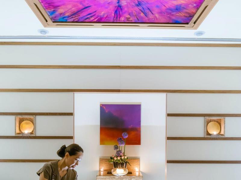 Žena dostává masáž horkými kameny v světlém, moderním wellness s barevným uměleckým dílem na stropě.