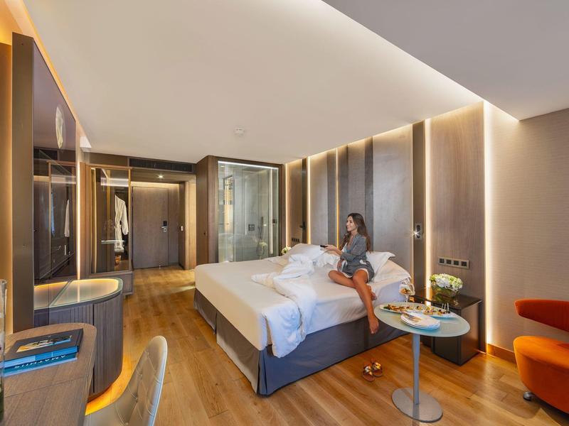 Moderní hotelový pokoj s manželskou postelí, dřevěnou podlahou a posezením s oranžovým křeslem.