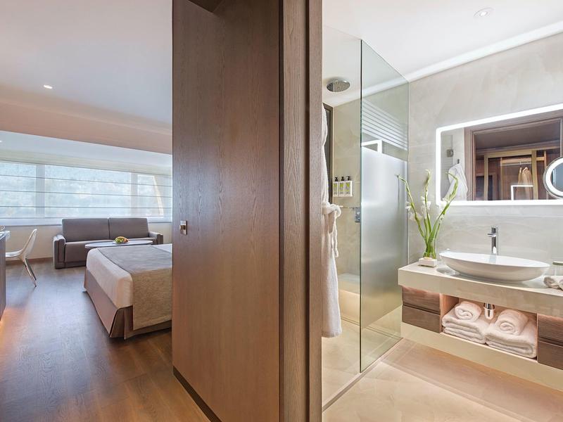Moderní hotelový pokoj se dřevěnou podlahou, manželskou postelí, posezením a světlou koupelnou se skleněnou sprchou.