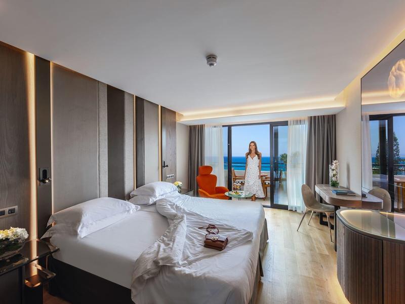 Moderní hotelový pokoj se dvěma postelemi, posezením a velkým oknem s výhledem na moře.