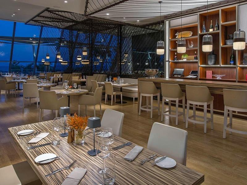 Moderní hotelová restaurace s dřevěnou podlahou, barem a velkými okny s nočním výhledem na město.