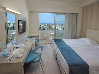 Hell eingerichtetes Hotelzimmer mit zwei Betten, Balkon, Schreibtisch und blauen Stühlen.