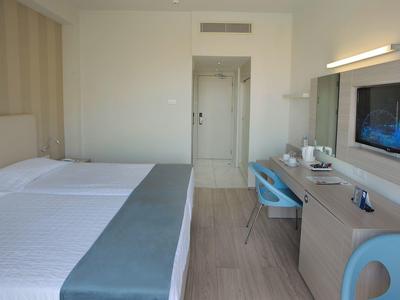 Helles Hotelzimmer mit zwei Betten, Schreibtisch, blauem Stuhl und Wandfernseher.