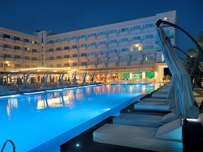 Evening view of an illuminated hotel pool with loungers and multi-story building in the background.