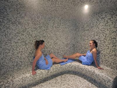 Zwei Frauen in blauen Handtüchern sitzen in einem dampfigen Mosaik-Dampfbad mit grauen Kacheln.