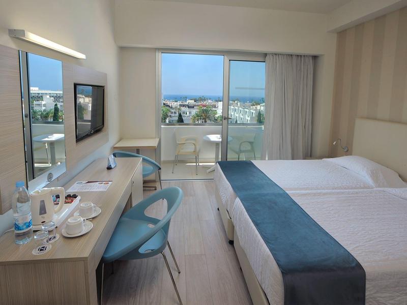 Hell eingerichtetes Hotelzimmer mit zwei Betten, Balkon, Schreibtisch und blauen Stühlen.