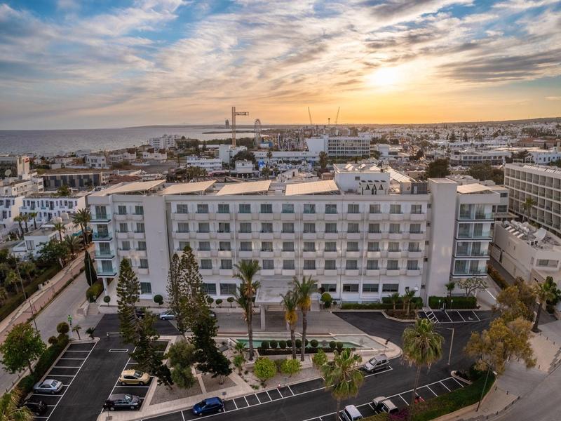 Großes weißes Gebäude mit mehreren Stockwerken, umgeben von Palmen und Autos bei Sonnenuntergang.