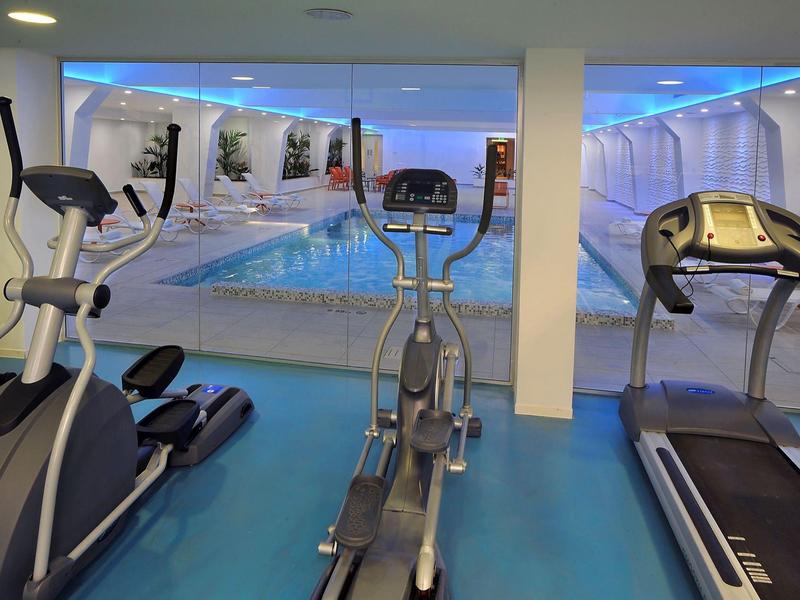 Fitnessraum mit Laufband, Crosstrainer und Fahrrad vor großem Schwimmbecken, moderne Beleuchtung.
