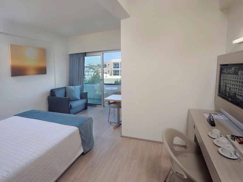 Helles Hotelzimmer mit Doppelbett, Stuhl, Schreibtisch, Fernseher und Balkon mit Tisch und Stühlen.