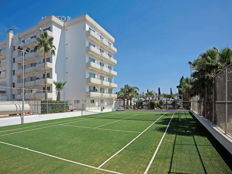 Moderner Mehrfamilienbau neben grünem Tennisplatz, umgeben von Palmen und blauem Himmel.
