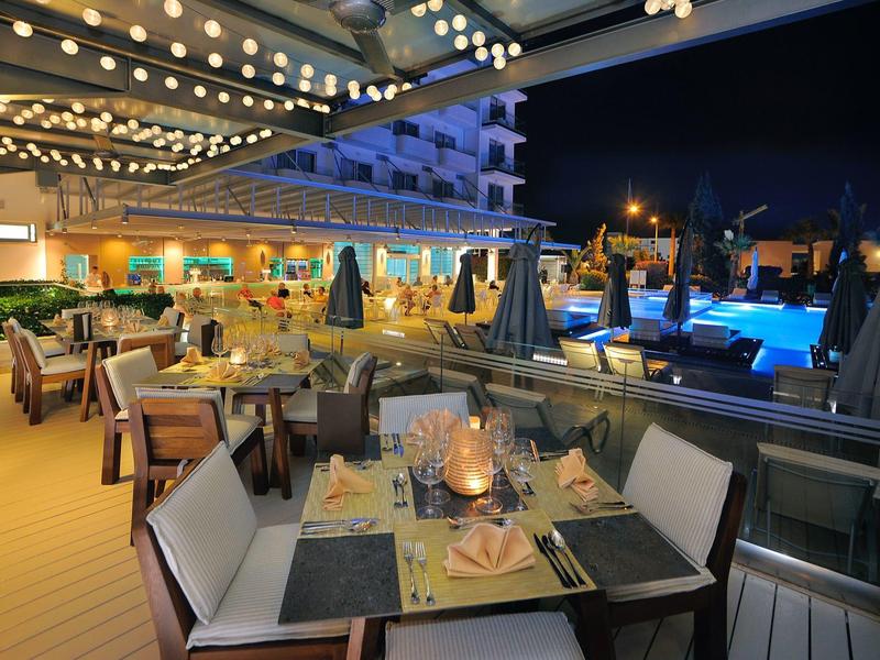 Elegantes Außenrestaurant bei Nacht mit gedeckten Tischen und beleuchtetem Pool im Hintergrund.