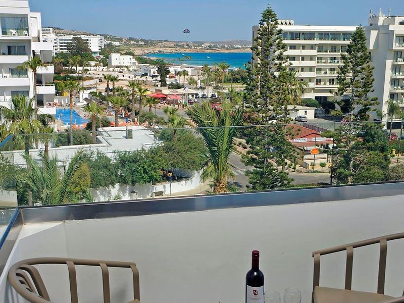 Balkon mit zwei Stühlen, kleinem Tisch, Obstschale und Weinflasche, im Hintergrund Hotels und Meer.