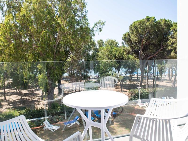 Balkon mit weißem Tisch und Stühlen, Glasgeländer, Bäume und sonniger Himmel im Hintergrund.