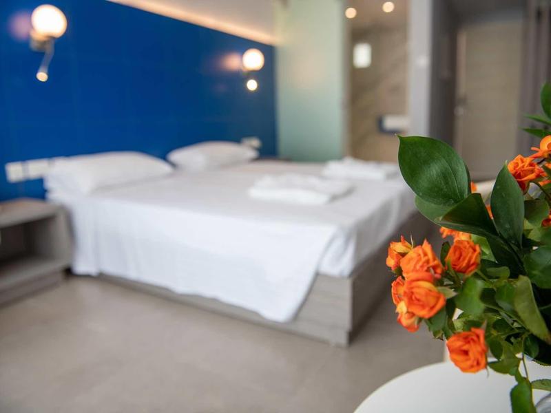 Ein weiß bezogenes Doppelbett vor dunkler Wand, im Vordergrund ein Tisch mit orangefarbenem Blumenstrauß.