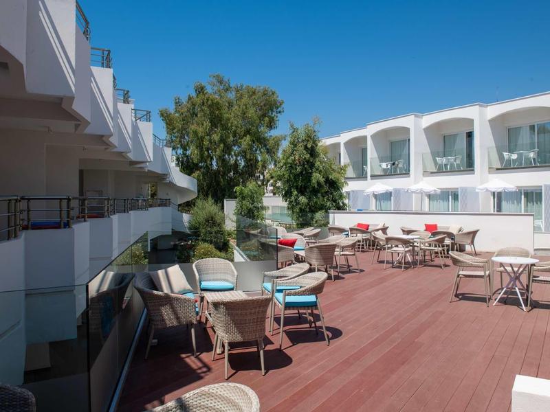 Große Terrasse mit weißen Tischen und Stühlen, zwei Gebäudeflügel, blauer Himmel und grüner Baum.