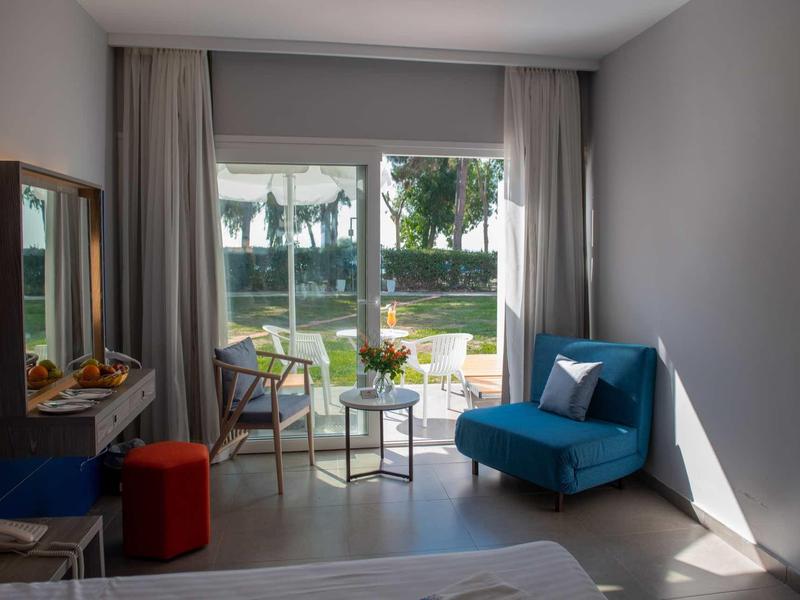 Helles Zimmer mit blauem Sessel, kleinen Tischen und zwei transparenten Stühlen vor Glasschiebetür.