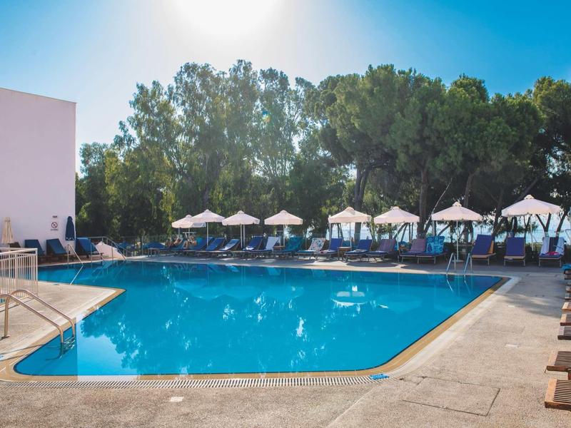 Großer blauer Pool mit Sonnenschirmen und Liegestühlen, umgeben von Bäumen und blauem Himmel.