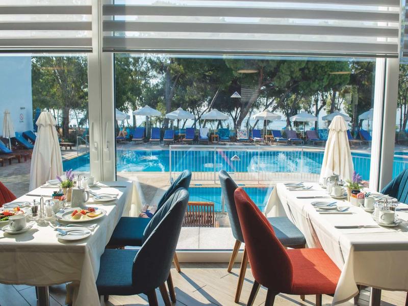 Restaurant mit gedeckten Tischen und Blick auf einen Pool mit Sonnenschirmen und Bäumen im Hintergrund.