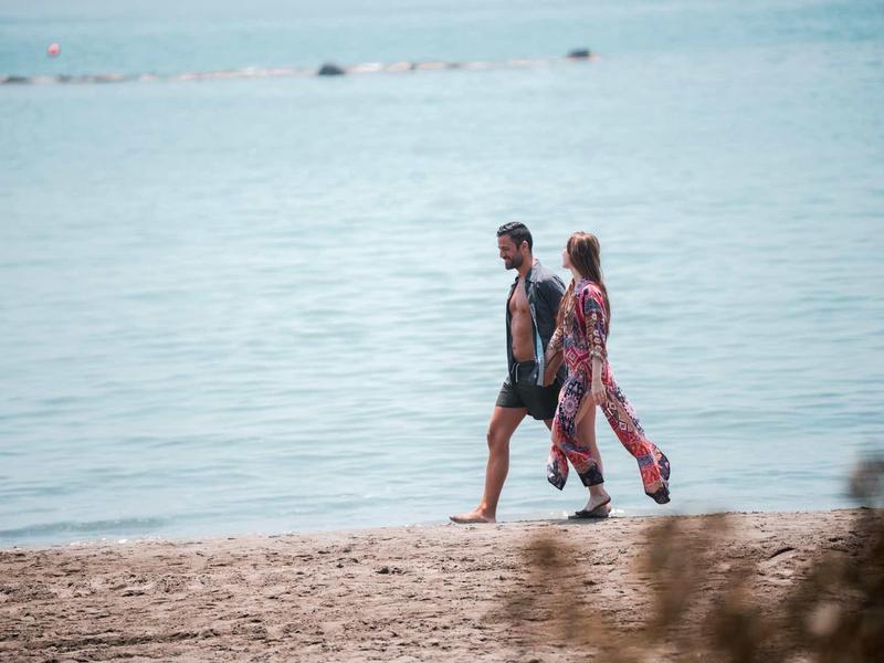 Zwei Menschen gehen barfuß am sandigen Strand entlang, im Hintergrund ruhiges Meer.