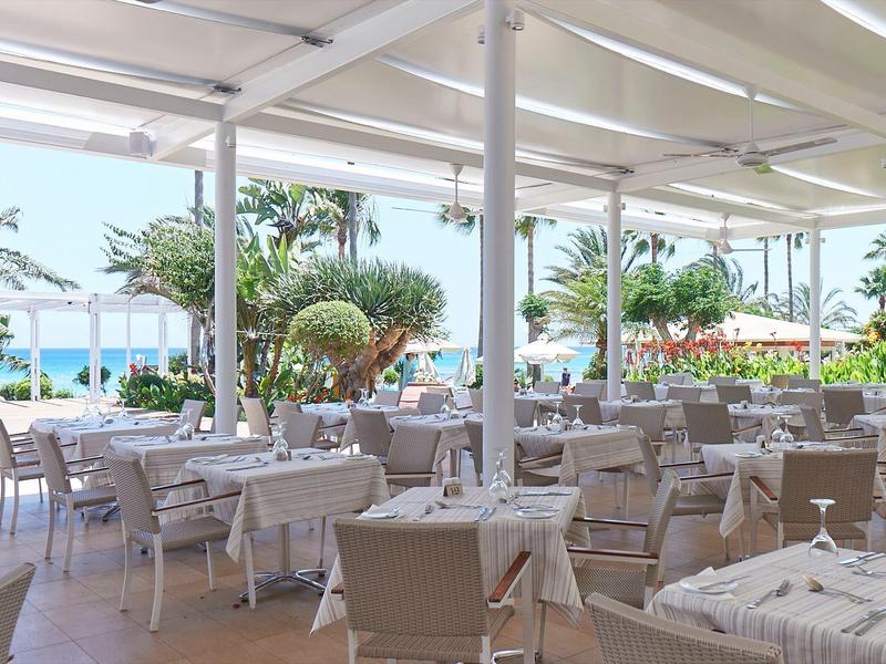 Terrassenrestaurant mit weißen Tischen und Stühlen, Blick auf Palmen und blaues Meer im Hintergrund.
