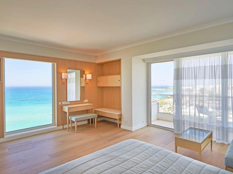 Helles Schlafzimmer mit großem Fenster, Blick auf das Meer, Holzmöbel und hellen Vorhängen.