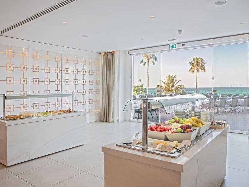 Heller Raum mit Buffetständern, frischem Obst und großem Fenster mit Meerblick und Palmen.