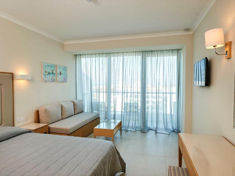 Helles Hotelzimmer mit Bett, Sofa, Tisch, Fernseher und Balkon mit Glas-Schiebetüren.