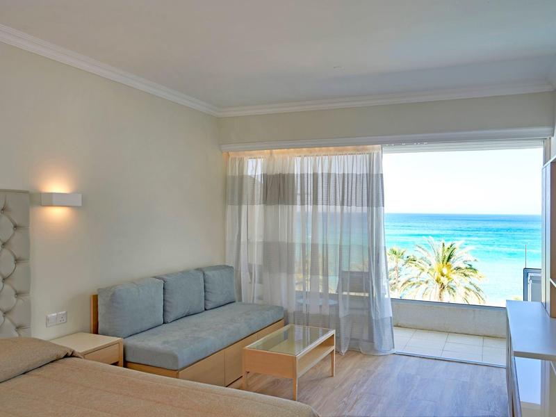 Helles Zimmer mit Sofa, Bett, Glas-Couchtisch, Balkon und Meerblick durch große Fenster.