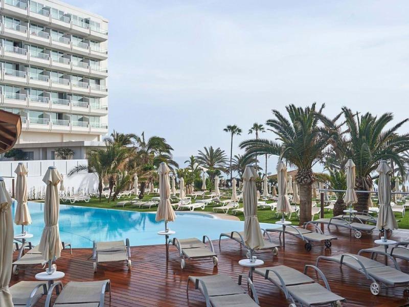 Hotelpoolbereich mit Liegestühlen, Sonnenschirmen, Palmen und einem mehrstöckigen Gebäude im Hintergrund.