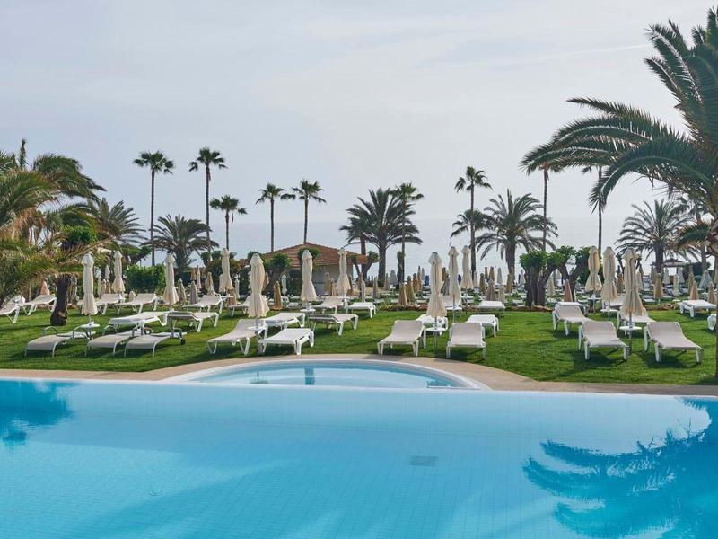 Blick auf einen Pool mit blauer Wasserfläche und Liegestühlen umgeben von Palmen und Rasen.