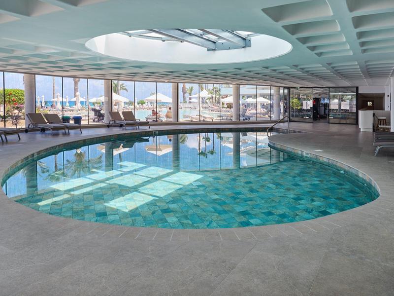 Runder Innenpool mit Glasdach und Blick auf die Stadt, umgeben von Liegestühlen.